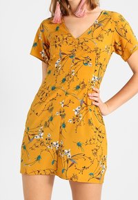 Sinapin keltainen lyhyt mekko, jossa on V-aukko, lyhyet hihat ja valkoisia, vihreitä sekä sinisiä kukkakuvioita. Sileä kangaspinta.