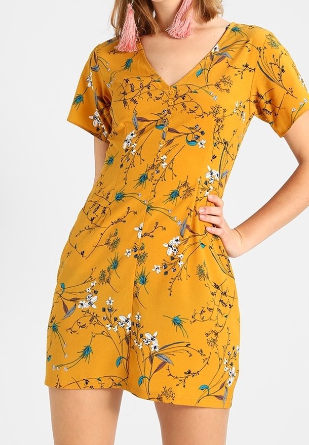 Sinapin keltainen lyhyt mekko, jossa on V-aukko, lyhyet hihat ja valkoisia, vihreitä sekä sinisiä kukkakuvioita. Sileä kangaspinta.