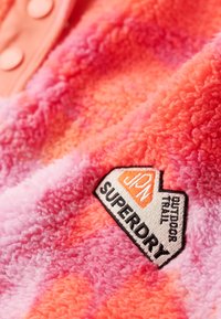 Blødt fleece-stof i gradient toner af pink og orange med et broderet Superdry-logo på nederste højre hjørne.
