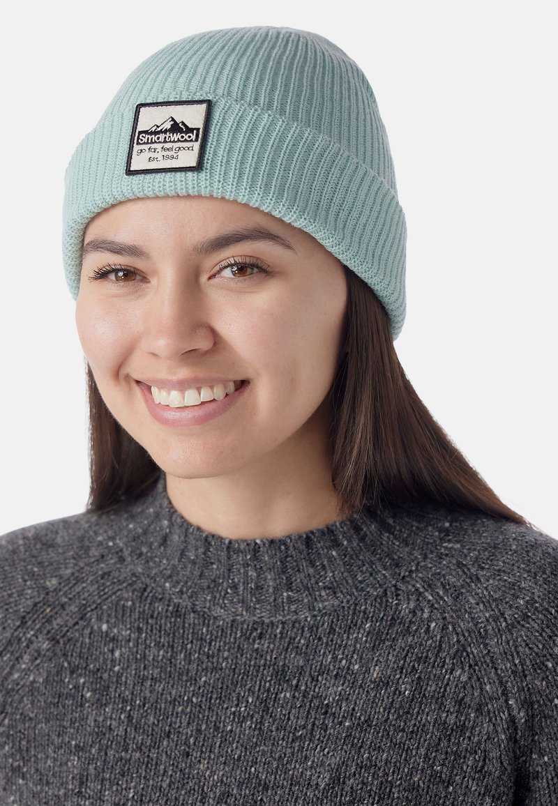 Gorro de punto acanalado azul claro con una etiqueta en blanco y negro, llevado sobre un suéter oscuro de textura gris con un cuello alto.