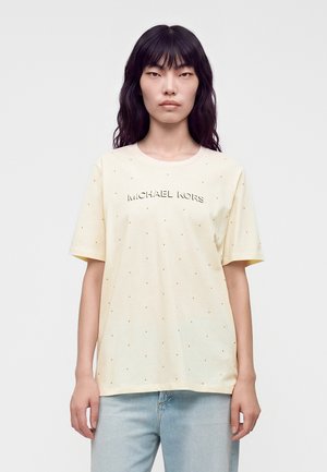 Kvinde med langt sort hår iført en lysegul Michael Kors t-shirt med små prikkede pynt og lyseblå jeans mod en hvid baggrund.