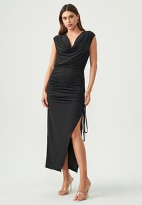 Schwarzes Kleid mit tiefem V-Ausschnitt und kurzen Ärmeln. Raffung am Taillenbereich und ein hoher Schlitz auf einer Seite. Glattes, elastisches Material.
