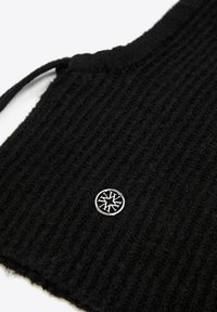 Canotta nera in lana coste con toppa tonda in argento con un logo geometrico nell'angolo inferiore. Testurizzata per il calore.