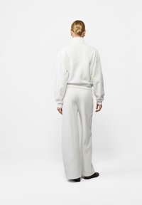 Kobieta z blond włosami związanymi w kok, ubrana w biały longsleeve i szerokie spodnie, stojąca na tle gładkiej białej ściany.