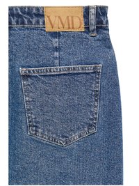 Vedere din spate a unei perechi de blugi din denim albastru, cu un buzunar dreptunghiular simplu și o etichetă din piele maro deschis cu inscripția „VMD” pe talie.