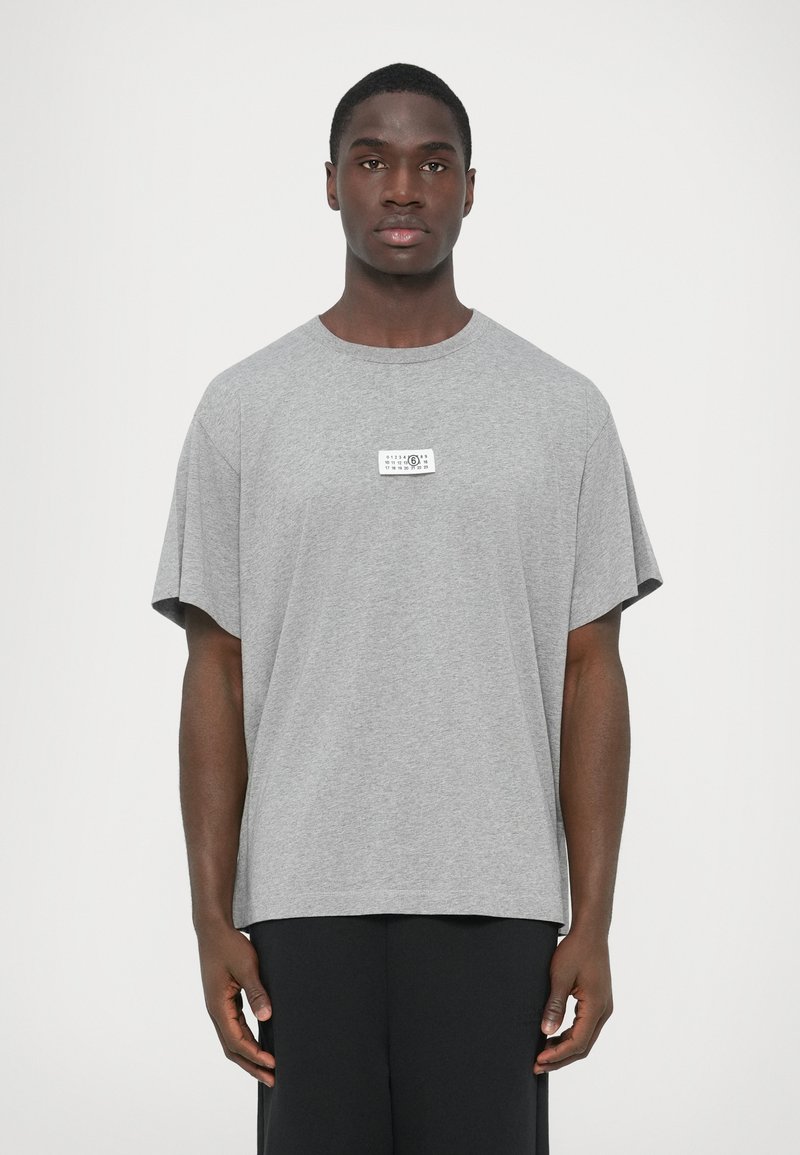 Siva bombažna majica s kratkimi rokavi, okroglim izrezom in pravokotno etiketo na sprednji strani. Ohlapen kroj z ravnim robom na dnu.