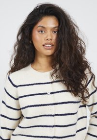 Giovane donna dai lunghi capelli ricci, che indossa un maglione bianco con righe orizzontali blu navy e bottoni bianchi, mentre guarda verso la camera.