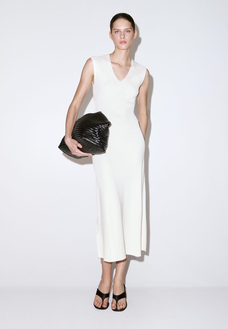Femme portant une robe midi blanche sans manches, tenant une grande pochette noire texturée et portant des talons ouverts noirs, debout devant un fond uni.