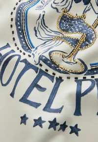 Tessuto bianco con testo stampato blu "HOTEL" e cinque stelle, con ricamo in perline dorate che delineano un motivo. Accenti blu aggiuntivi visibili.