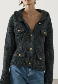 Graue Strickjacke mit Kragen, verziert mit goldfarbenen Knöpfen und zwei Fronttaschen, zeigt ein strukturiertes, grob gestricktes Design.