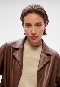 Casaco em pele sintética castanha com gola dentada e detalhes em botão, combinado com um suéter bege. O cabelo curto do modelo está estilizado.