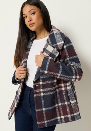 Femme portant une chemise blanche et un pantalon bleu avec un manteau à carreaux violet, bleu et blanc, tenant les revers contre un fond uni.
