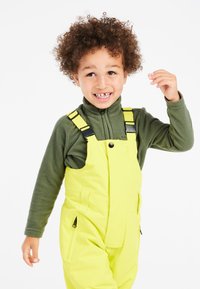 Lyse gule overalls med sort hardware og lynlåslommer, båret over en mørkegrøn fleece. Barn smilende med krøllet hår, hånd løftet.