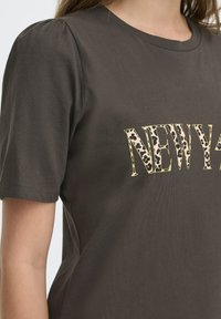 Kobieta ubrana w ciemno-brązowy t-shirt z krótkimi rękawami, na którym dużymi literami w panterkę znajduje się napis "NEWY" na wysokości klatki piersiowej.