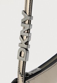 Τσάντα DKNY με μεταλλικό λουρί που λάμπει και ανάγλυφα γράμματα, λεία μαύρη ύλη και κλείσιμο με φερμουάρ με ασημέν ιες μεταλλικές λεπτομέρειες.