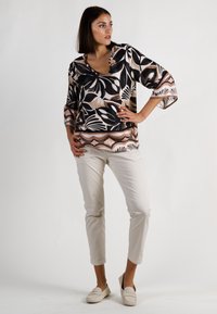 Bluse mit schwarzen und beigen Blattmustern, runder Ausschnitt, 3/4-Ärmel; kombiniert mit cremefarbenen Hosen und hellen, lässigen Schuhen.
