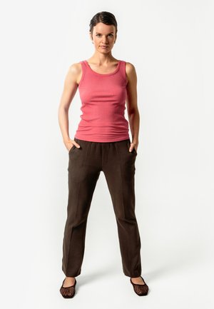 Frau steht mit den Händen in den Taschen, trägt ein ärmelloses pinkes Oberteil, braune Hosen und schwarze Mesh-Ballerinas vor weißem Hintergrund.