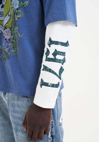 Kurzärmliges blaues T-Shirt mit grafischem Design; lange weiße Ärmel mit grüner, abgenutzter Schrift, die "1871" liest. Getragen mit hellblauen, abgenutzten Jeans.