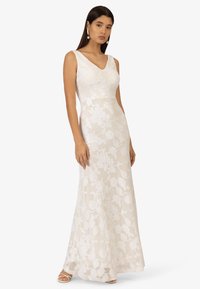 Weißes Kleid mit floraler Spitzenüberlagerung, V-Ausschnitt und tailliertem Oberteil. Fällt bis zum Boden mit einem durchsichtigen Stoff. Einfaches, elegantes Design.