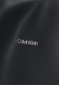Borsa a mano in pelle nera testurizzata con logo "CalvinKlein" in argento. La superficie presenta una sottile trama a grana.