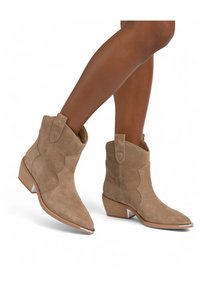 Botas de tobillo de ante beige con puntera afilada, detalles de costura y tiradores. Presentan un tacón bajo apilado con un acento metálico.