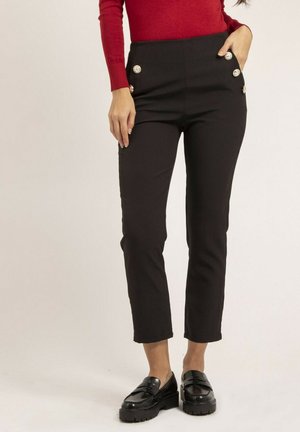Femme portant un pantalon noir taille slim avec des boutons dorés et des mocassins noirs, associée à un haut rouge à manches longues, debout devant un fond uni.