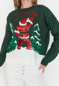 Zielony sweter dzianinowy z cekinowym wzorem Mikołaja, czerwonym napisem "HO HO HO" i białymi akcentami u dołu. Wygodny krój, świąteczny motyw.