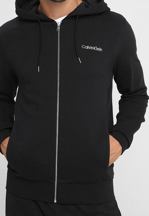 Mann trägt schwarzen Calvin Klein Zipper-Hoodie mit Händen in den Taschen, weißes T-Shirt darunter, und Kordeln hängen von der Kapuze.