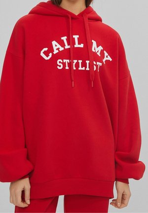 Osoba nosząca za dużą czerwoną bluzę z kapturem z białym napisem "CALL MY STYLIST" oraz pasujące czerwone spodnie na jednolitym jasnym tle.