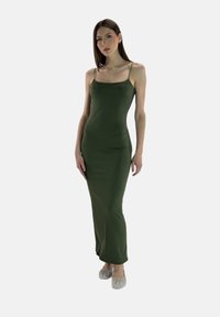 Groene, nauwsluitende spaghetti-riem jurk gemaakt van gladde stof, met een rechte halslijn en een lengte tot op de vloer, gecombineerd met zilveren platte schoenen.