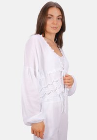 Weiße Baumwollbluse mit lässiger Passform, gewelltem Spitzenbesatz und Bindeschluss. Sie verfügt über lange Ärmel mit elastischen Bündchen und strukturierter Stoff.