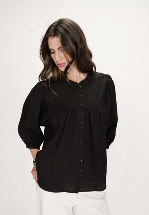 Femme aux longs cheveux noirs portant un chemisier noir ample à boutons avec des manches bouffantes et un pantalon blanc, regardant vers le bas les mains dans les poches.