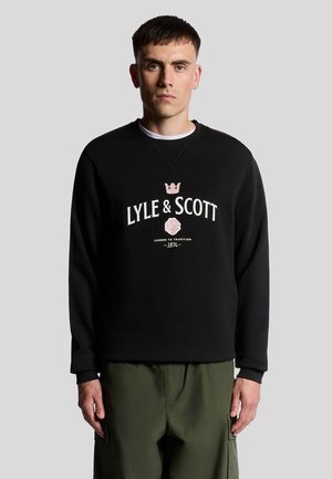 Schwarzer Sweatshirt mit Rundhalsausschnitt, versehen mit dem Lyle & Scott-Logo in Weiß und Pink, getragen mit olivgrünen Cargo-Hosen.