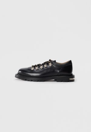 Black leather low-cut shoe with thick sole, metal eyelets, decorative silver charms on laces, and textured tread on the sole.