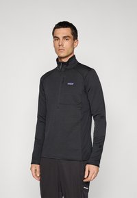 Man wearing a black long-sleeve half-zip fleece top with a Patagonia logo on the chest, standing against a plain light background.
