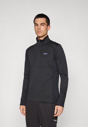 Man wearing a black long-sleeve half-zip fleece top with a Patagonia logo on the chest, standing against a plain light background.