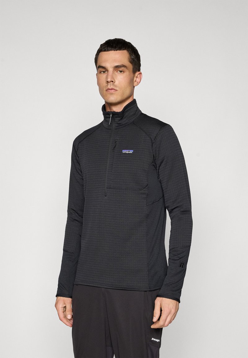 Man wearing a black long-sleeve half-zip fleece top with a Patagonia logo on the chest, standing against a plain light background.