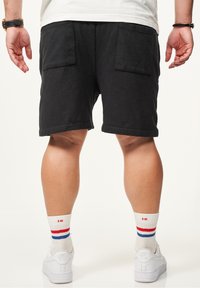 Schwarze Baumwollshorts mit zwei hinteren Taschen, kombiniert mit weißen Sneakers und gestreiften Knöchelsocken, von hinten betrachtet.