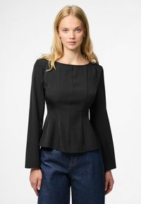 Top peplum nero con scollatura rotonda, maniche lunghe e cuciture verticali, abbinato a jeans blu a gamba larga, posizionato su uno sfondo neutro.