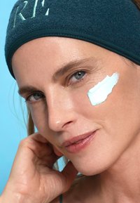 Jasnoniebieska kremowa substancja nałożona na policzek; modelka nosi ciemnoniebieską, futrzaną opaskę na głowę z białym haftem; gładka skóra, miękkie oświetlenie, niebieskie tło.