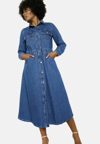 Džinsu krekla kleita ar garām piedurknēm, divām krūštura kabatām, pogu priekšpusi, elastīgu jostasvietu un līdz ceļiem garumu, vidēji zila mazgāšanas krāsā.