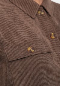 Brązowa koszula z żebrowanej kordury z guzikami w kolorze muszli oraz klapą na kieszeni na piersi.