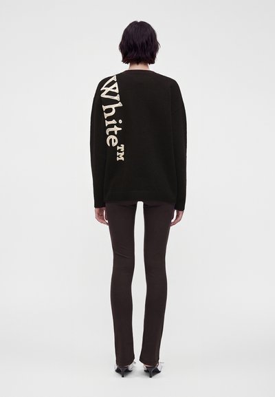 Personne aux cheveux courts et foncés, debout de dos, portant un pull noir avec un grand texte vertical "White" dans le dos et un pantalon slim sombre.