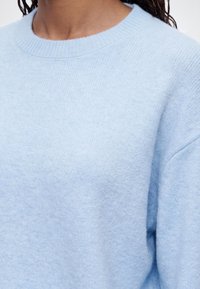 Personne portant un pull tricoté bleu clair à col rond avec un tissu texturé et des manches longues visibles sur l'épaule et le haut de la poitrine.