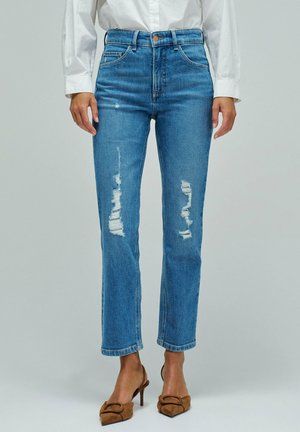 Mujer con vaqueros azules desgastados de cintura alta y piernas cortadas, combinados con tacones marrones de punta puntiaguda y una camisa blanca de manga larga.