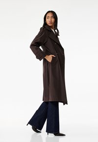 Gabardina marrón con grandes solapas, botones de doble botonadura, bolsillos laterales y cintura ceñida con cinturón, combinada con jeans oscuros de campana y tacones.