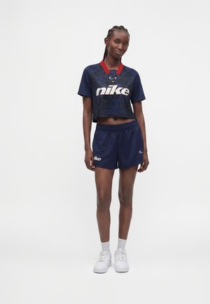 Γυναίκα στέκεται φορώντας σκούρο μπλε crop top Nike με κόκκινο γιακά και ταιριαστά σορτς, λευκές κάλτσες και λευκά αθλητικά σε απλό φόντο.