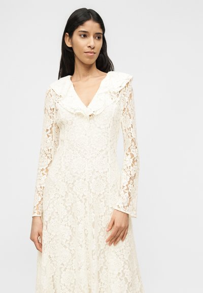 Γυναίκα με μακρυμάνικ ο φόρεμα από κρέμ δαντέλα με λουλουδένια σχέδια και με γιακά με βολάν, που στέκεται μπροστά σε ένα απλό φωτεινό φόντο.