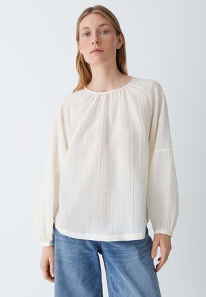 Vrouw met lang blond haar, gekleed in een losse witte blouse met textuur en blauwe spijkerbroek, tegen een effen achtergrond.