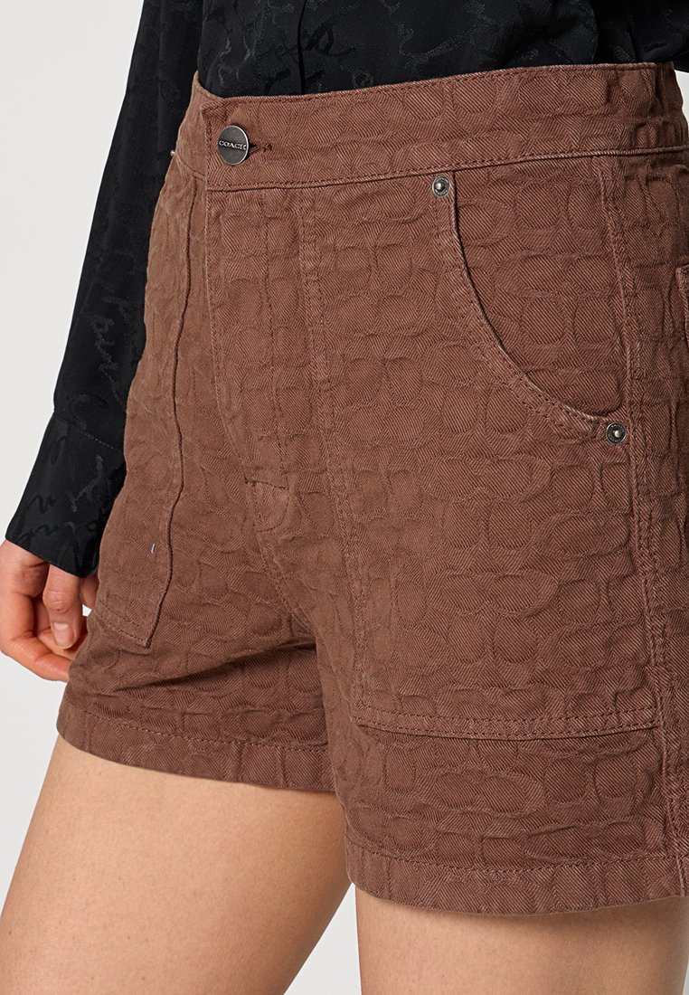 Braune Shorts mit Struktur, die ein geometrisches Muster aufweisen, zwei frontale Taschen und einen Metallknopfverschluss haben. Das Material scheint leicht und langlebig zu sein.